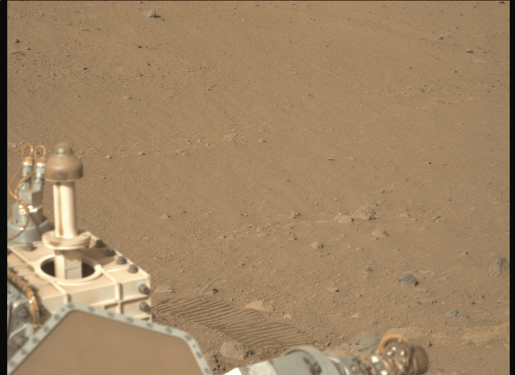 Color photo across the dusty, reddish-brown floor of Jezero Crater on Mars, with part of Perseverance's deck and a fresh wheel track visible in the foreground.
