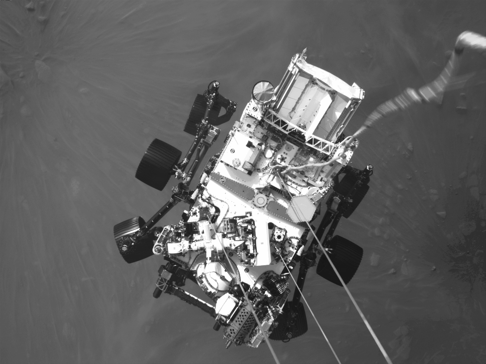 Black-and-white overhead view of NASA's Perseverance rover suspended on cables beneath its rocket-powered descent stage moments before landing on Mars.