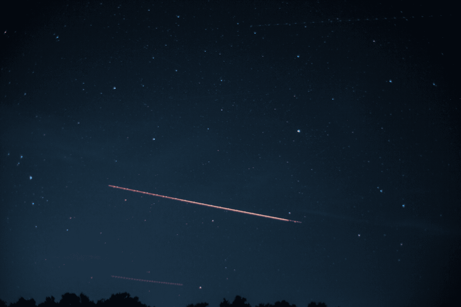 Star field looking east with a segmented red aircraft trail and a dark treeline along the bottom