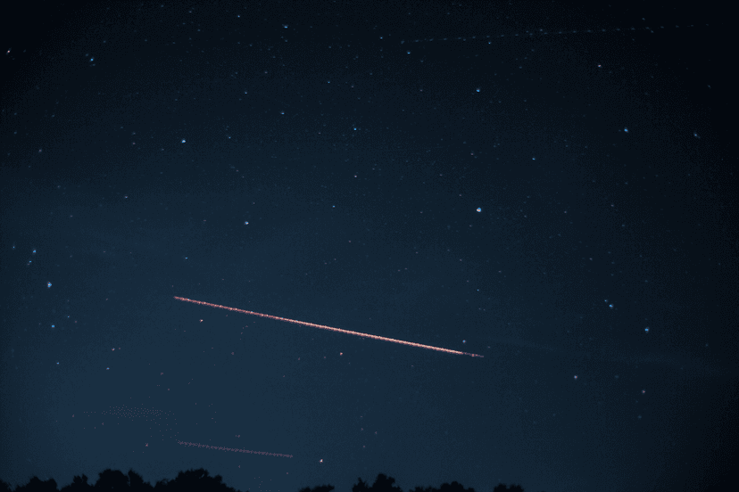 Star field looking east with a segmented red aircraft trail and a dark treeline along the bottom